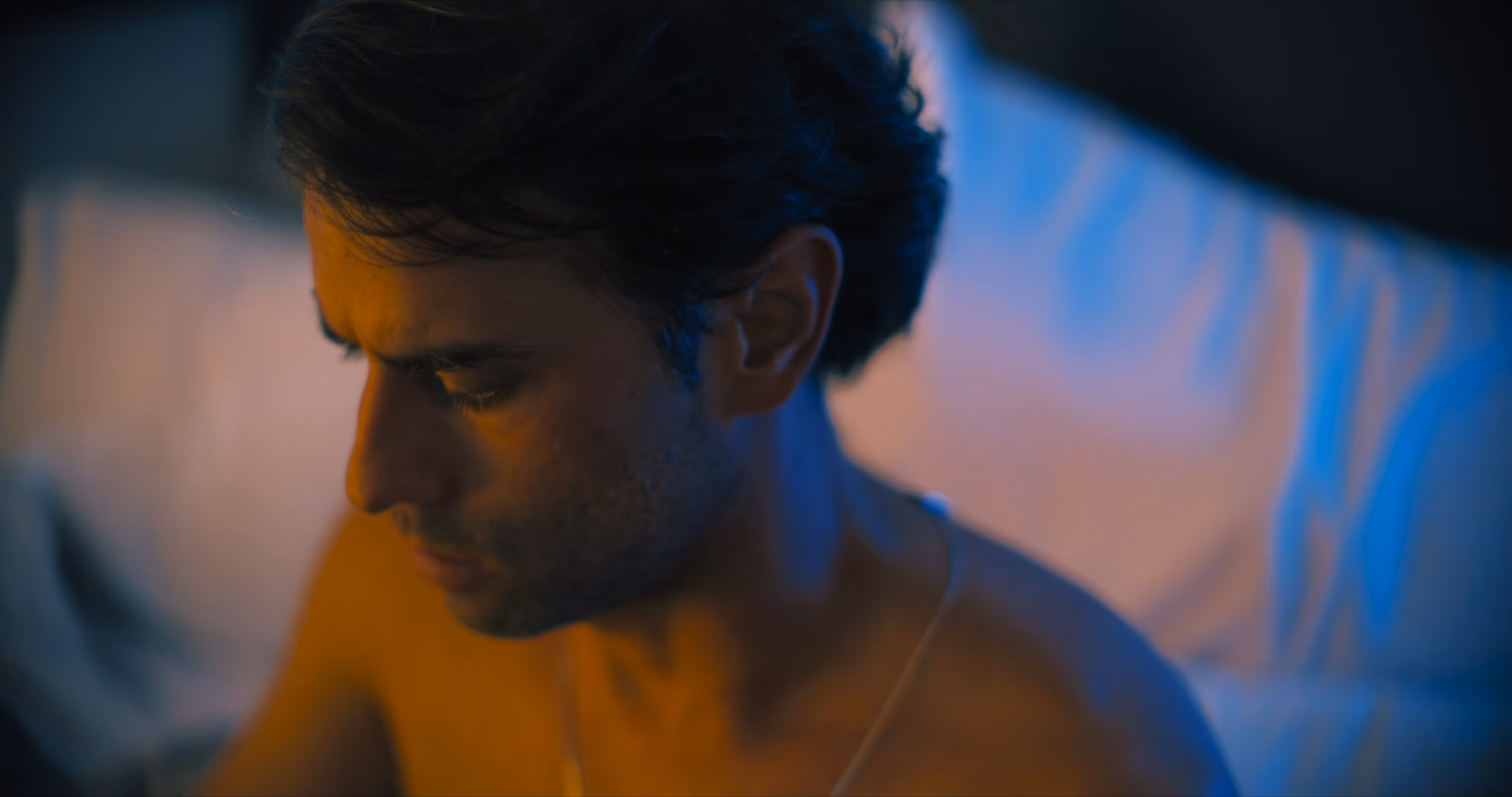 Side profile of a shirtless man with short dark hair sitting on a bed illuminated by warm and blue lighting.