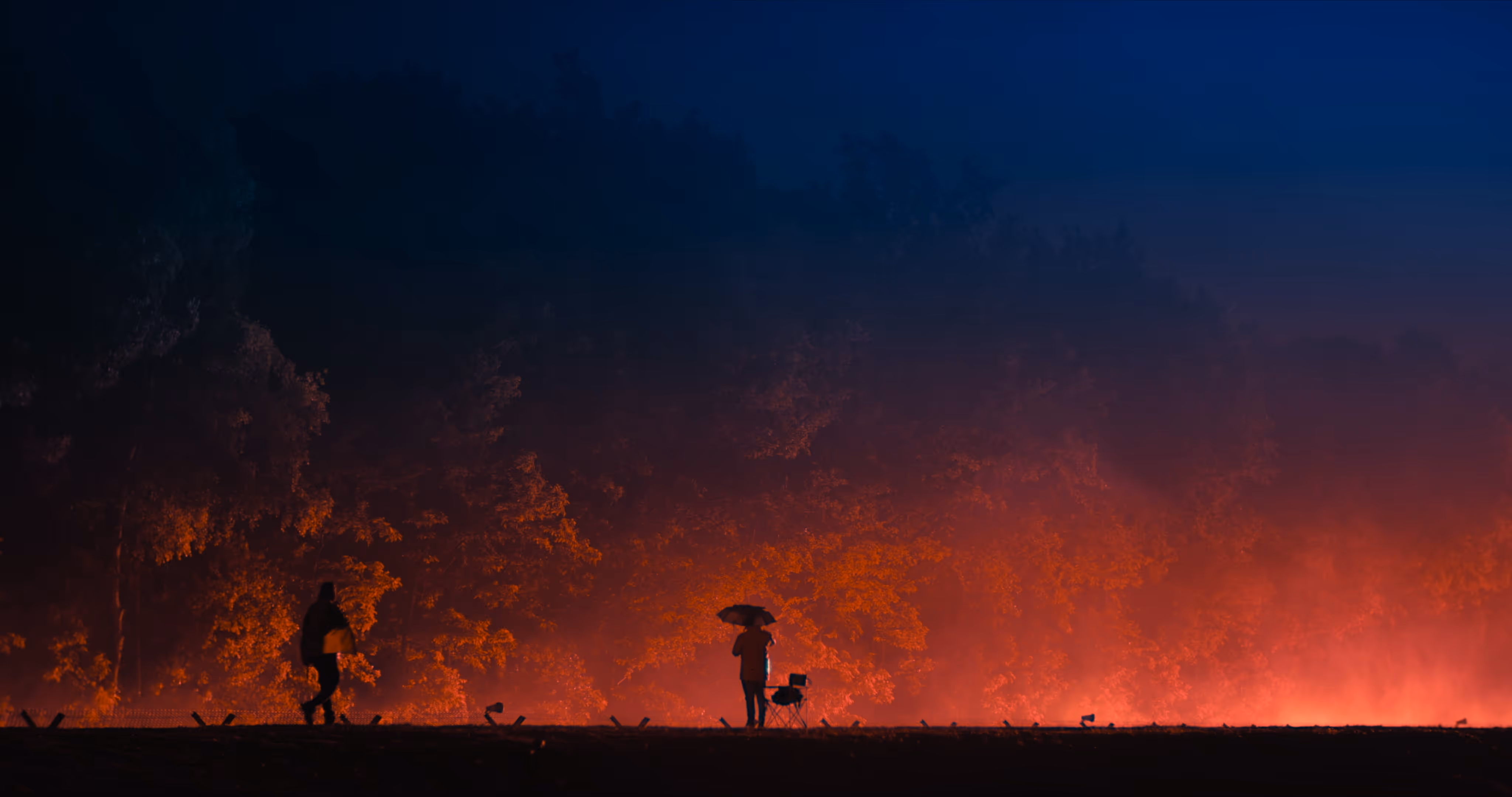Two people silhouetted against glowing orange light with trees in the background during night, one holding an umbrella.