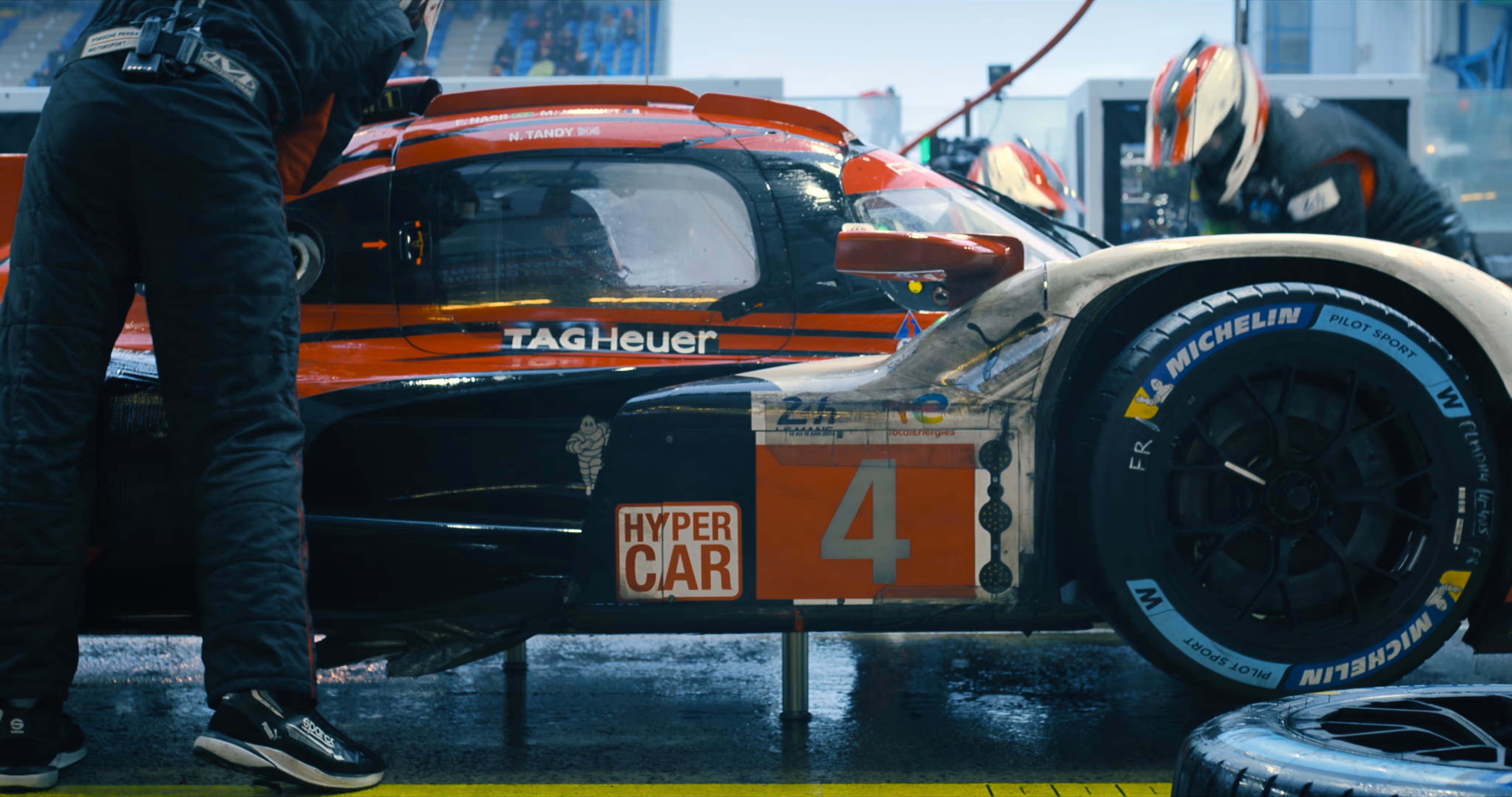 Side view of a race car numbered 4 in a pit stop with team members working and Michelin Pilot Sport tires.