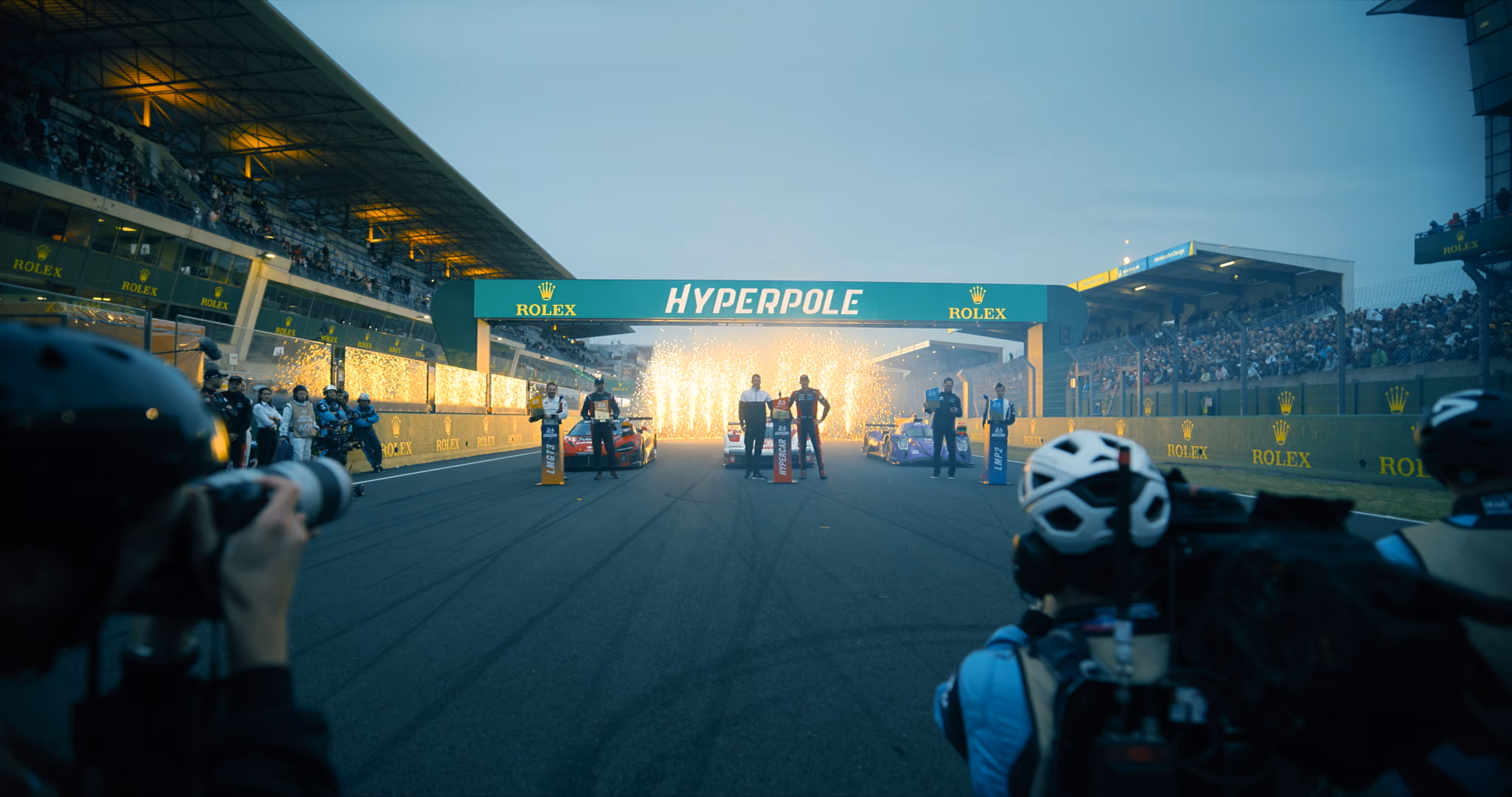 Race drivers standing on a track under a 'HYPERPOLE' sign with fireworks going off behind them and photographers capturing the scene.