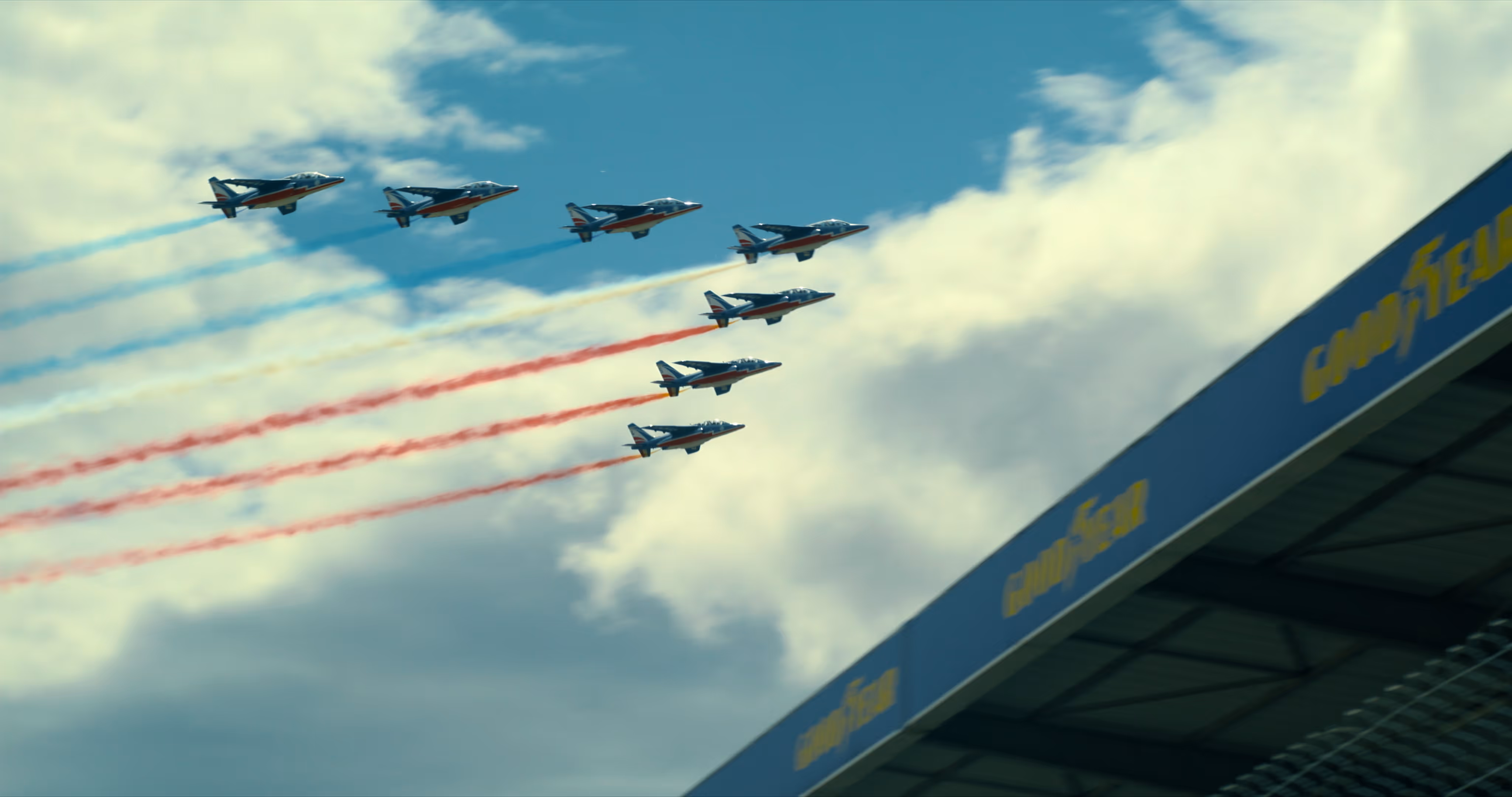 Eight aircraft flying in formation with trails of red, yellow, and blue smoke against a partly cloudy sky near a grandstand with Goodyear branding.