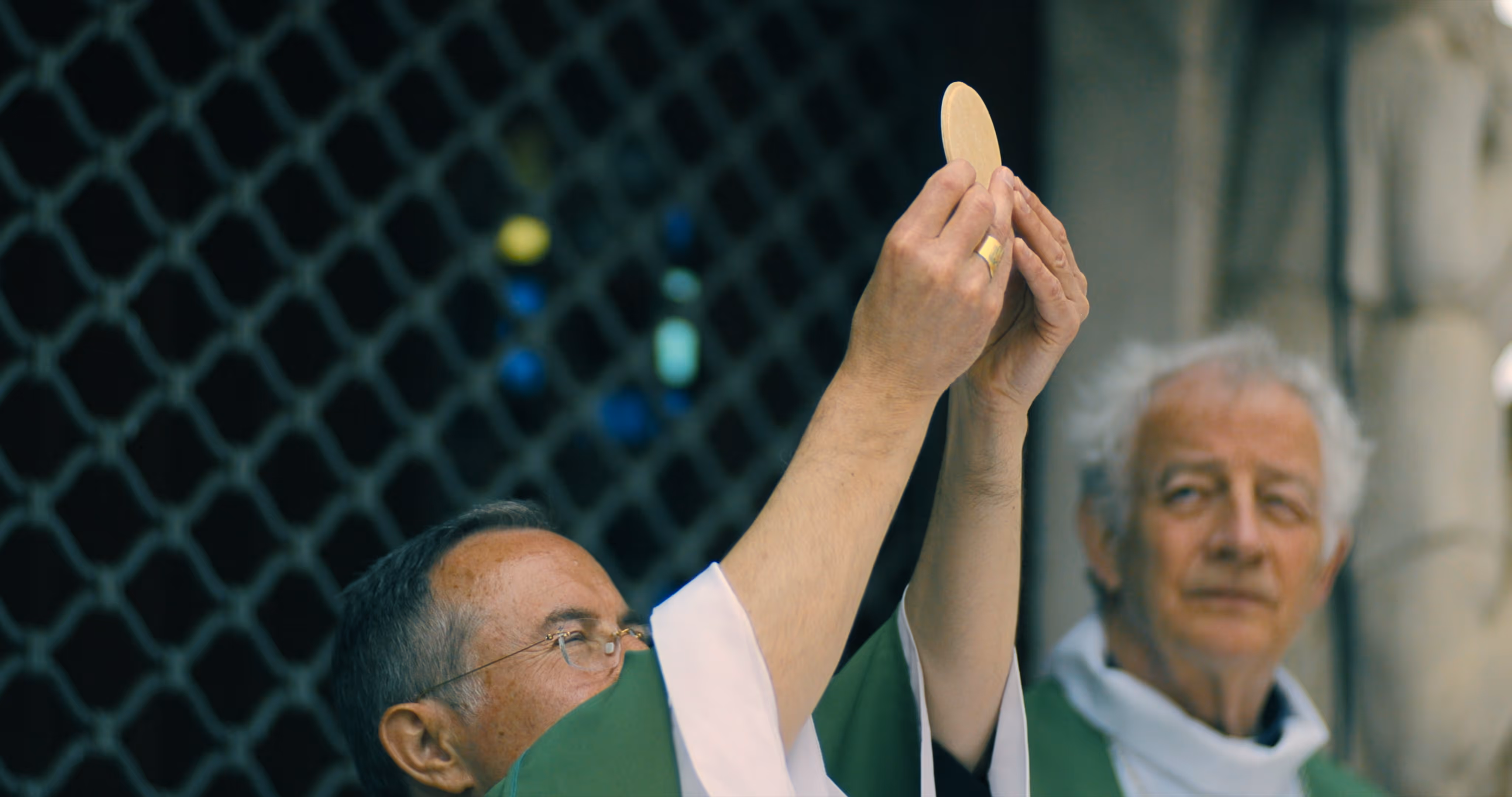 Catholic priest raising a communion wafer during a religious ceremony with another clergyman in the background.