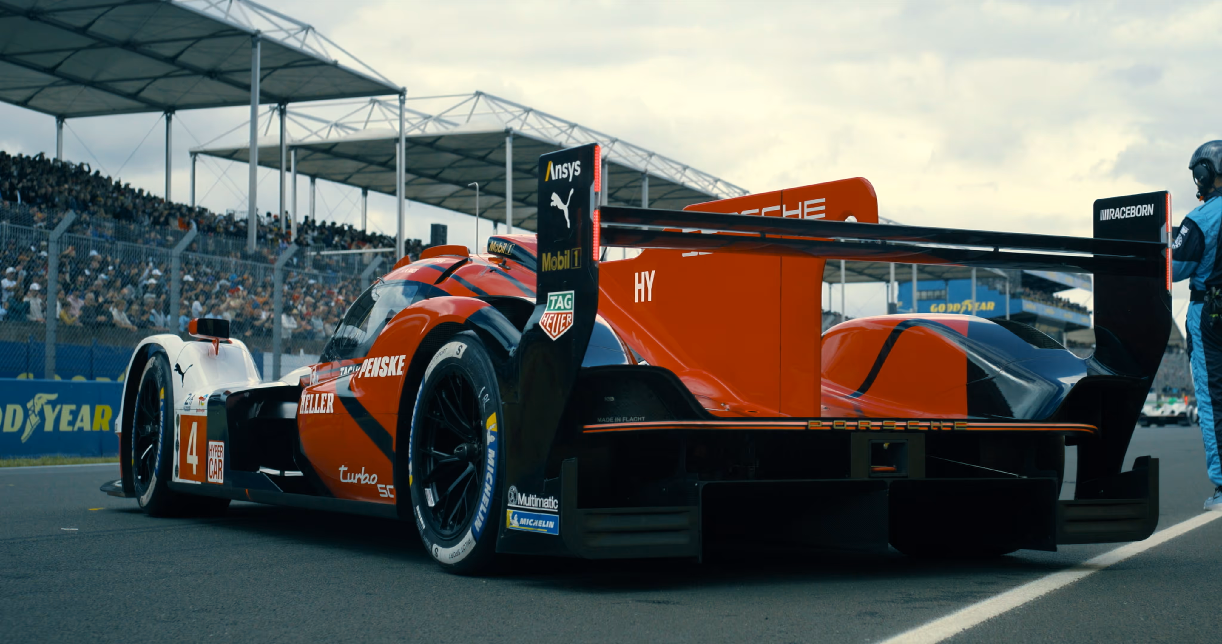 Red and white Porsche race car with large rear wing on racetrack, crowds and pit crew visible.