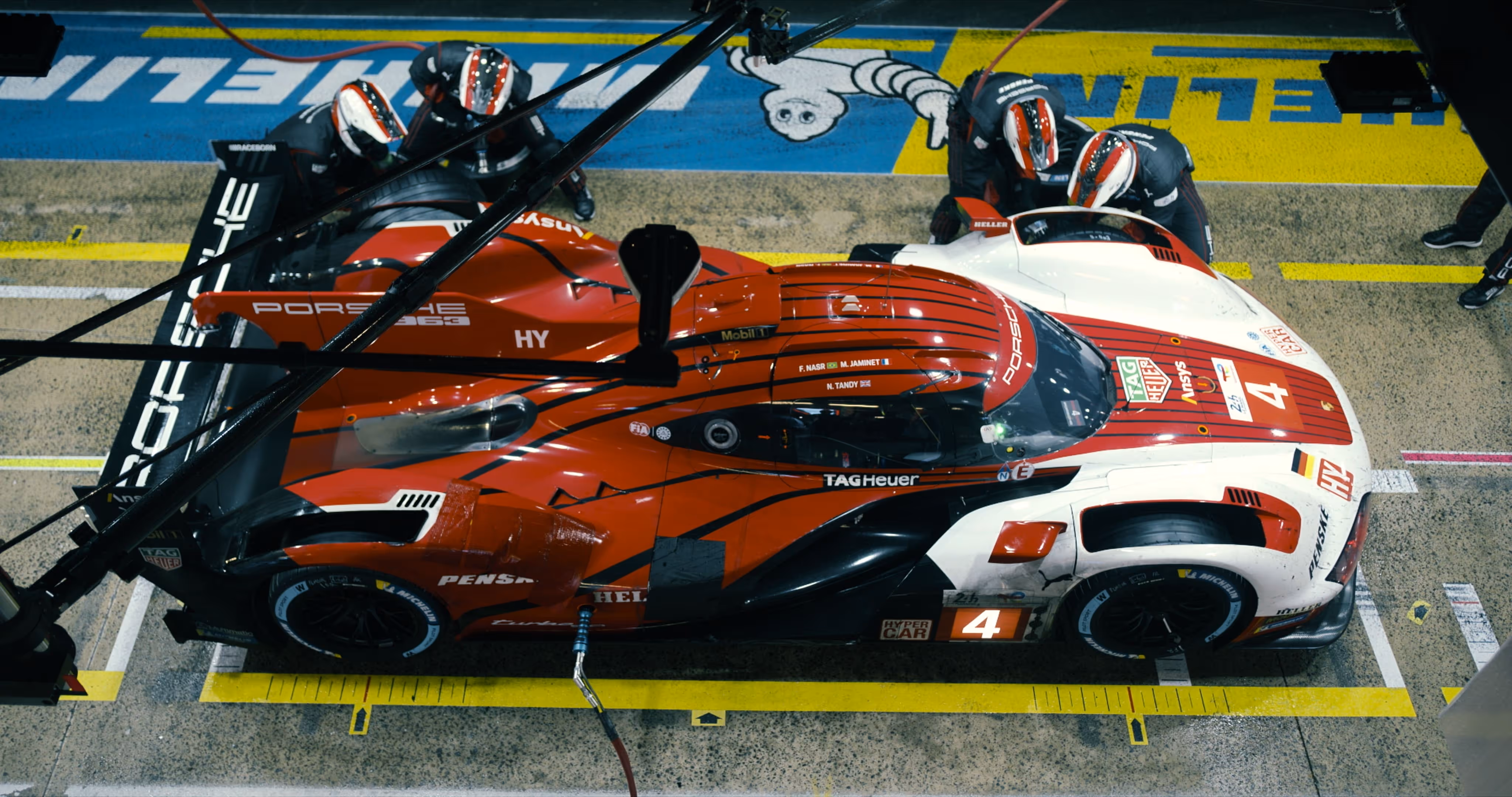 Top view of a red and white Porsche race car number 4 in a pit stop with team members servicing it.