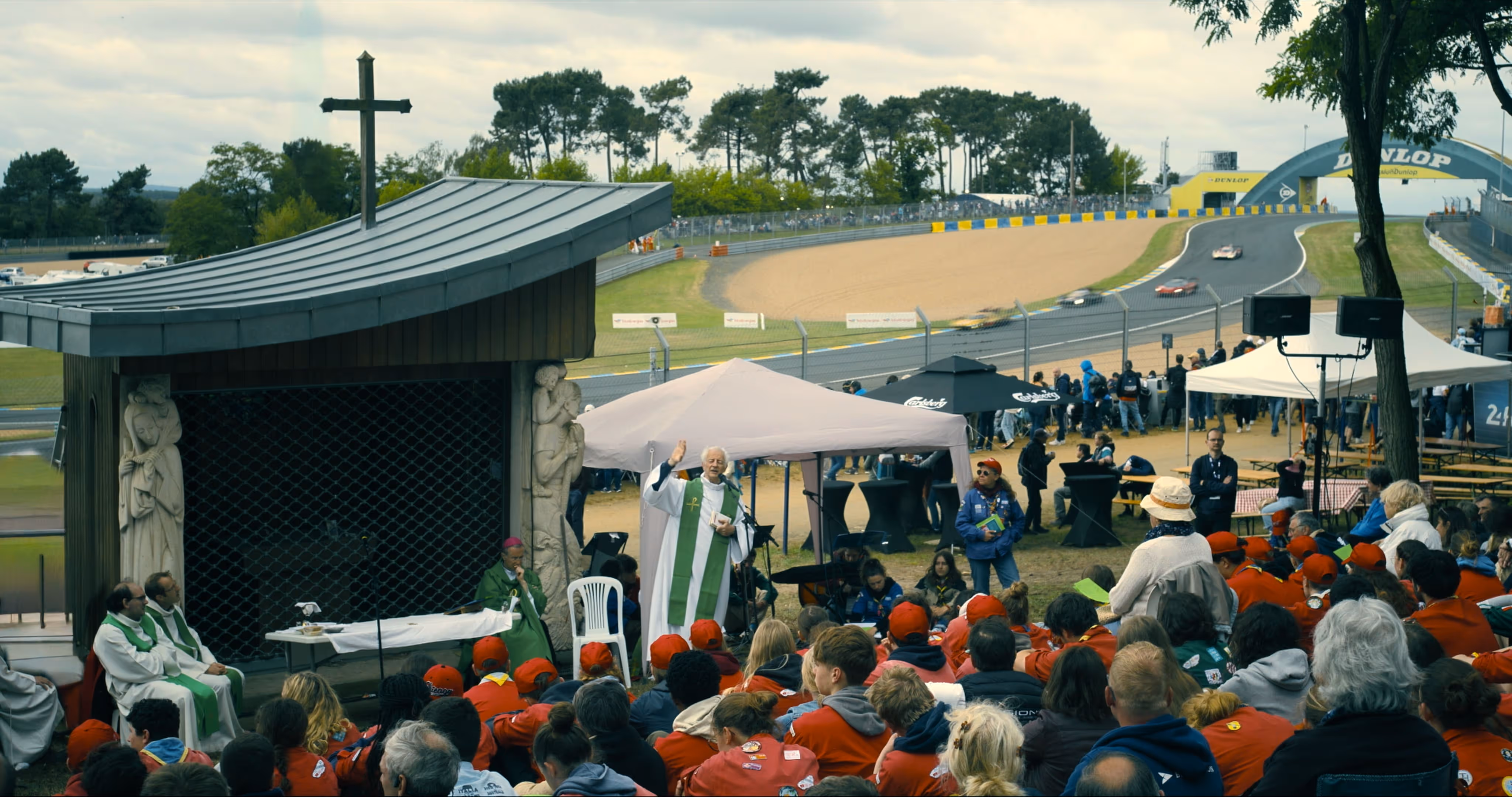Outdoor religious service with a priest speaking to a seated crowd near a race track with cars passing by Dunlop bridge.