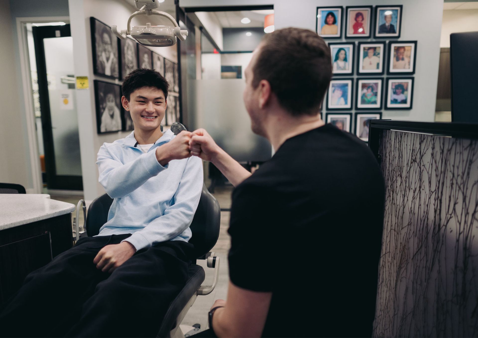 Smiling young man sitting in a dental chair fist bumping with a man in a black shirt in a dental office.