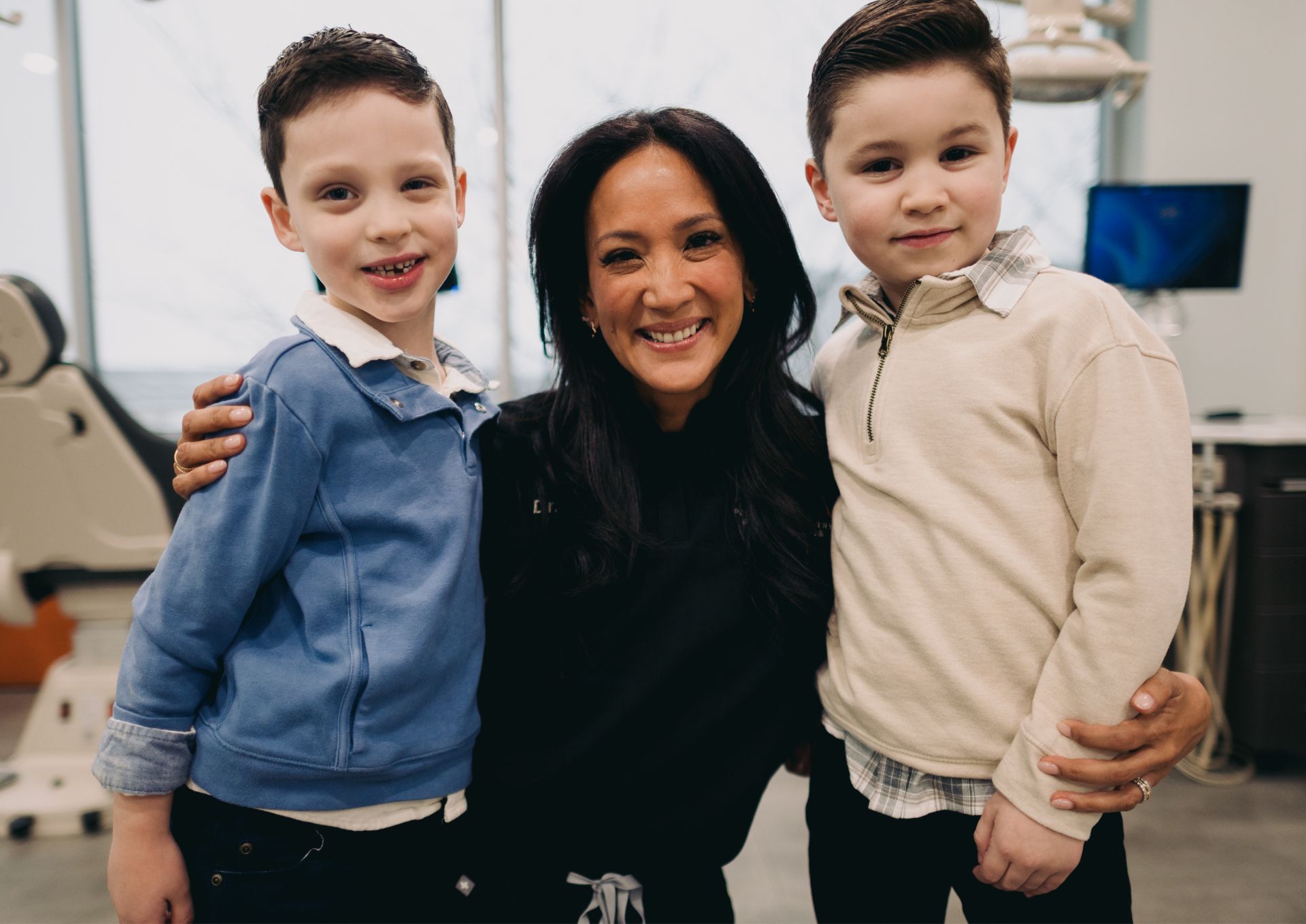 Smiling woman with long dark hair poses between two young boys in a modern medical or dental office.