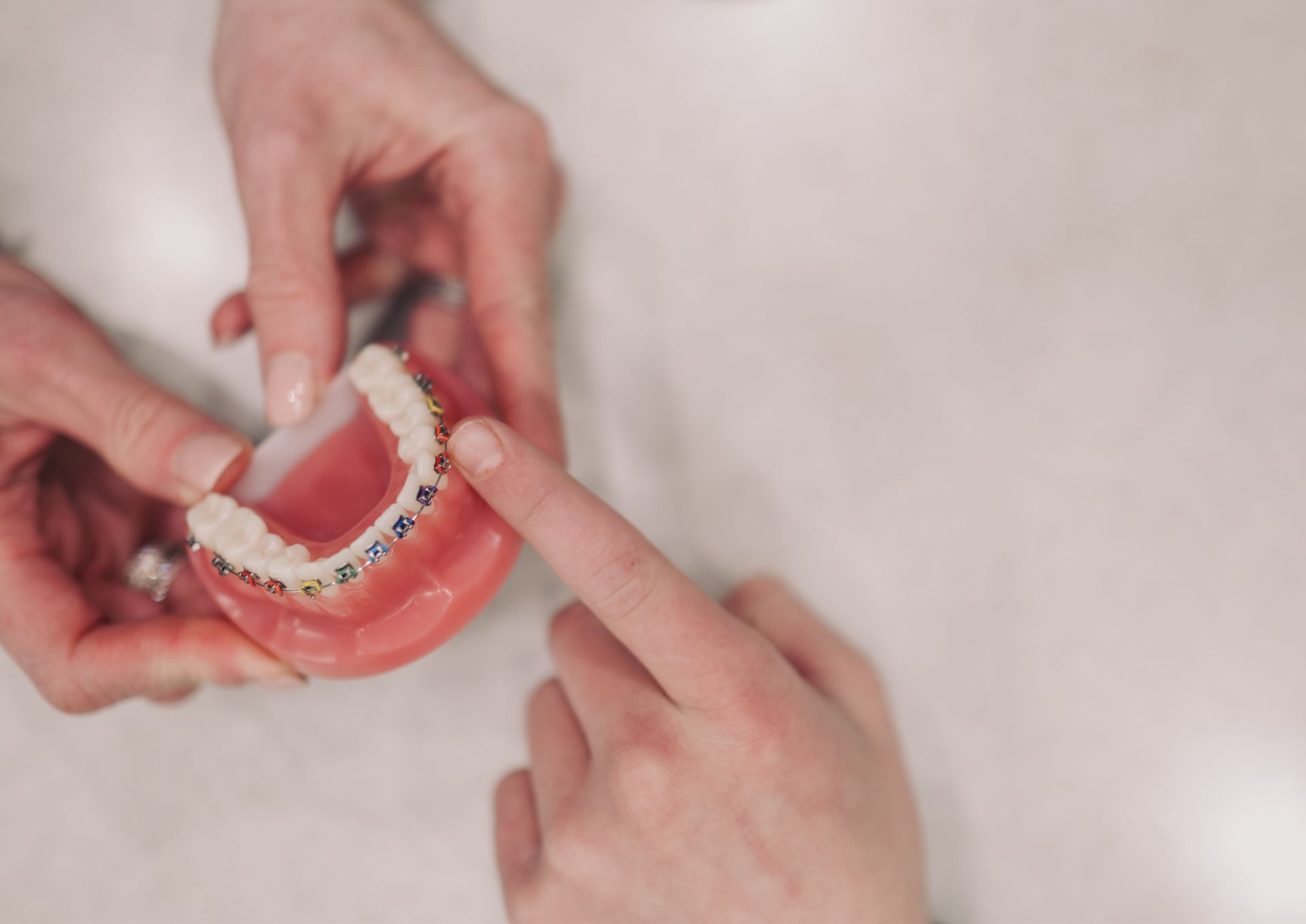 Hands holding a dental model showing colorful braces on teeth while a finger points at them.