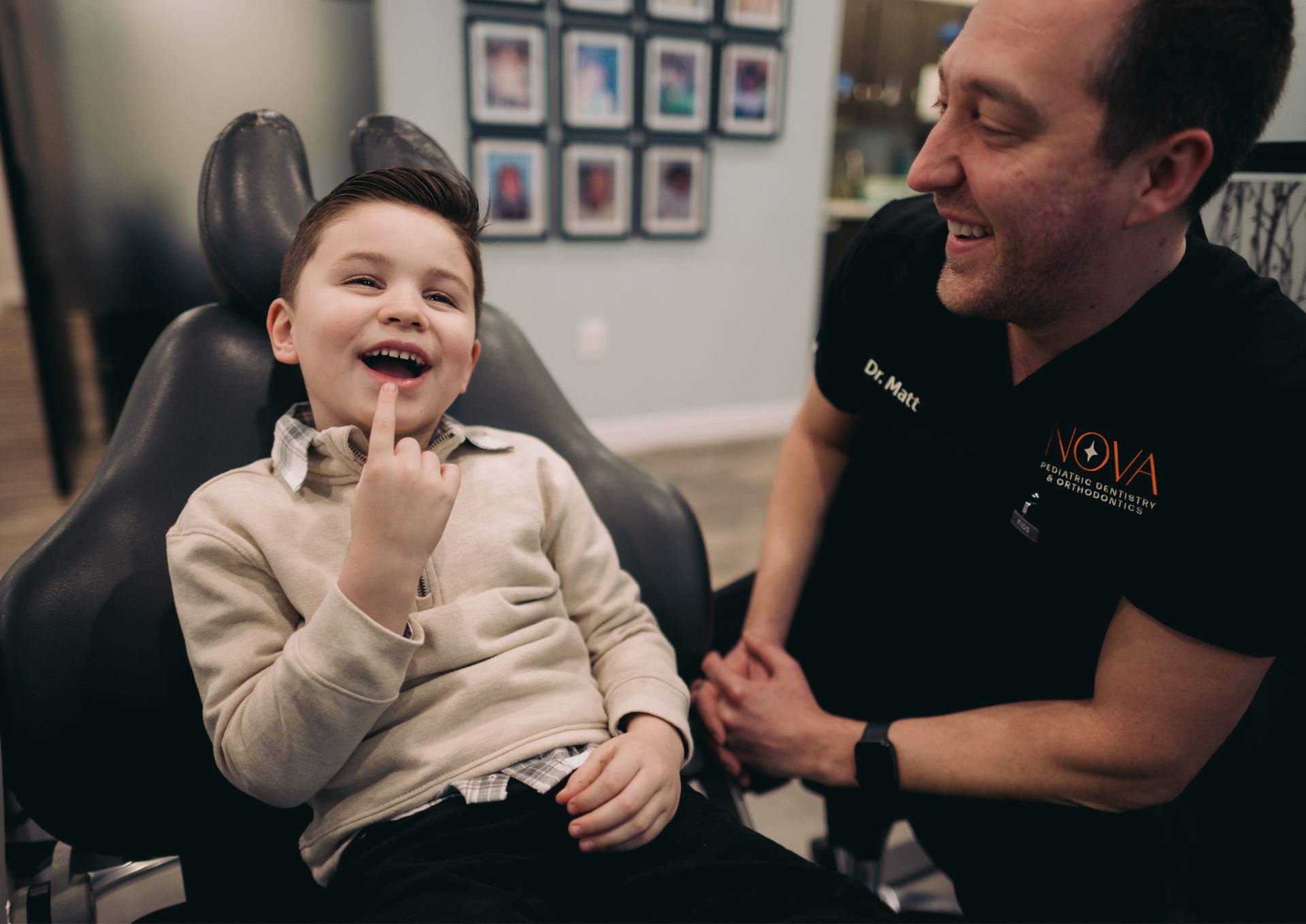 Smiling boy sitting in a dental chair pointing at his mouth with a male dentist in black scrubs beside him laughing.