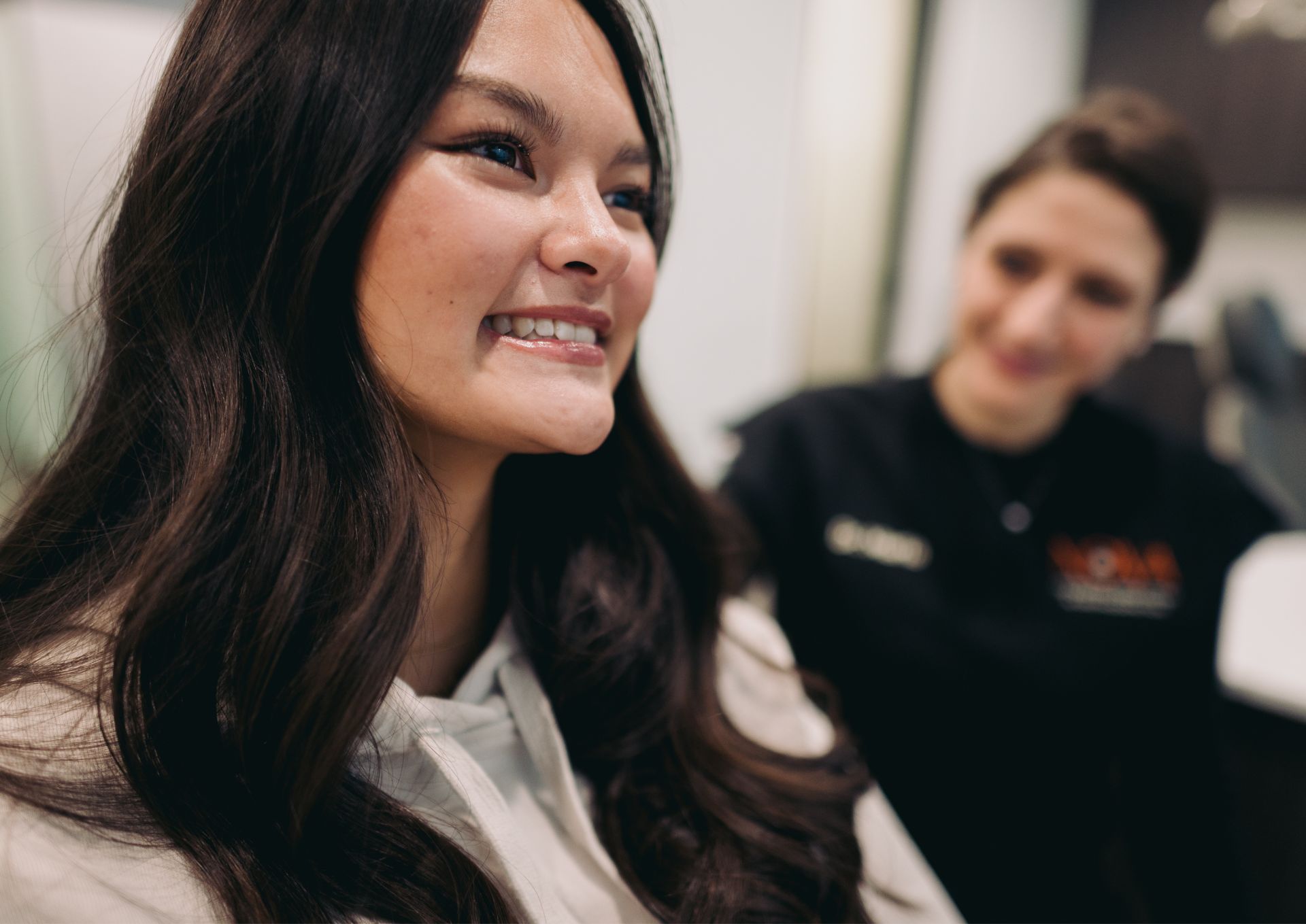Smiling young woman with long dark hair, with a blurred person in medical scrubs in the background.
