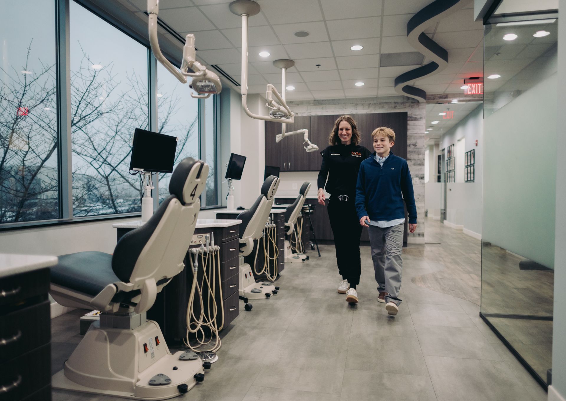 Orthodontic clinic with a female dentist walking alongside a smiling young boy in the treatment area with dental chairs and equipment.