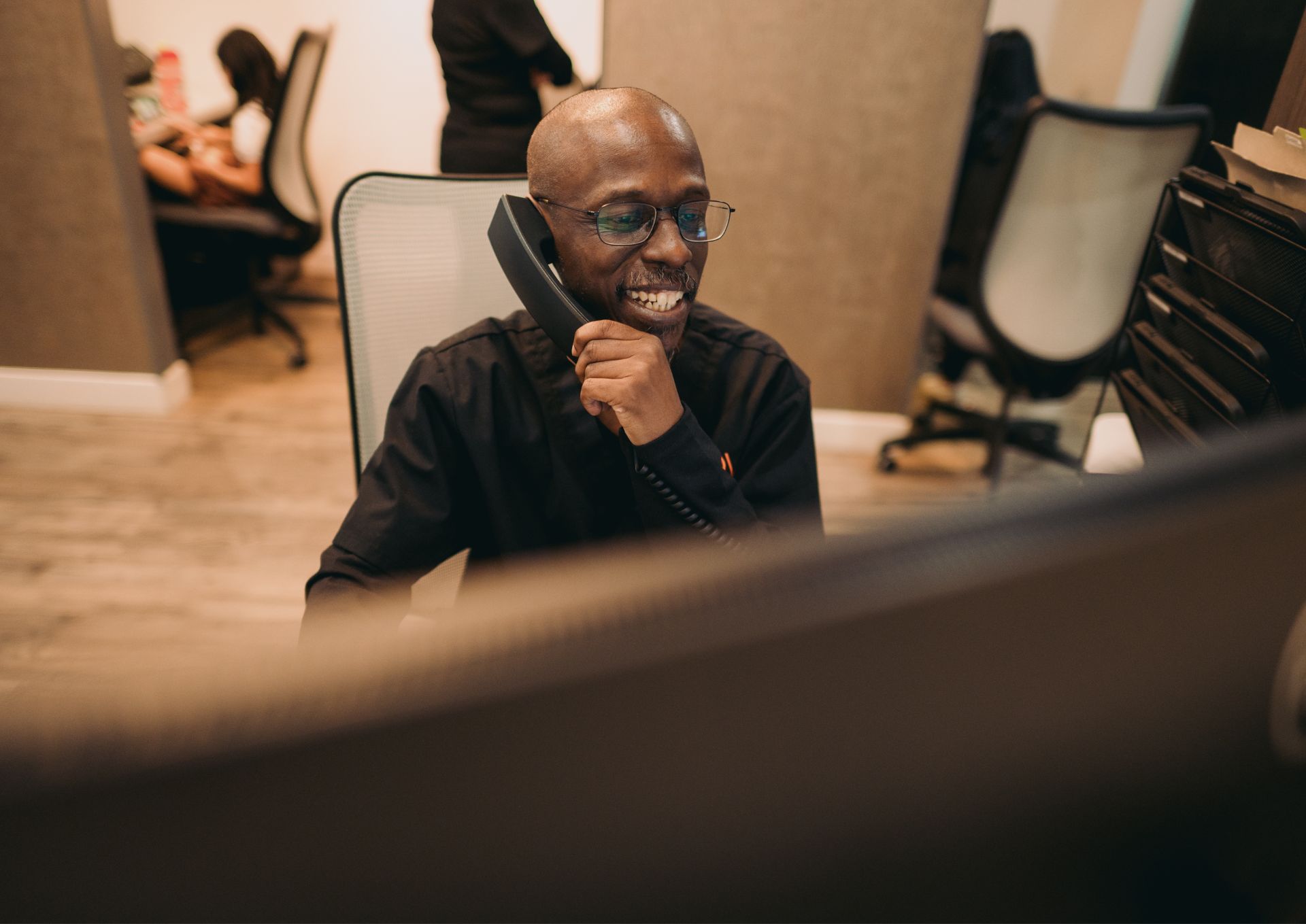 Smiling man wearing glasses talking on a desk phone in an office setting.