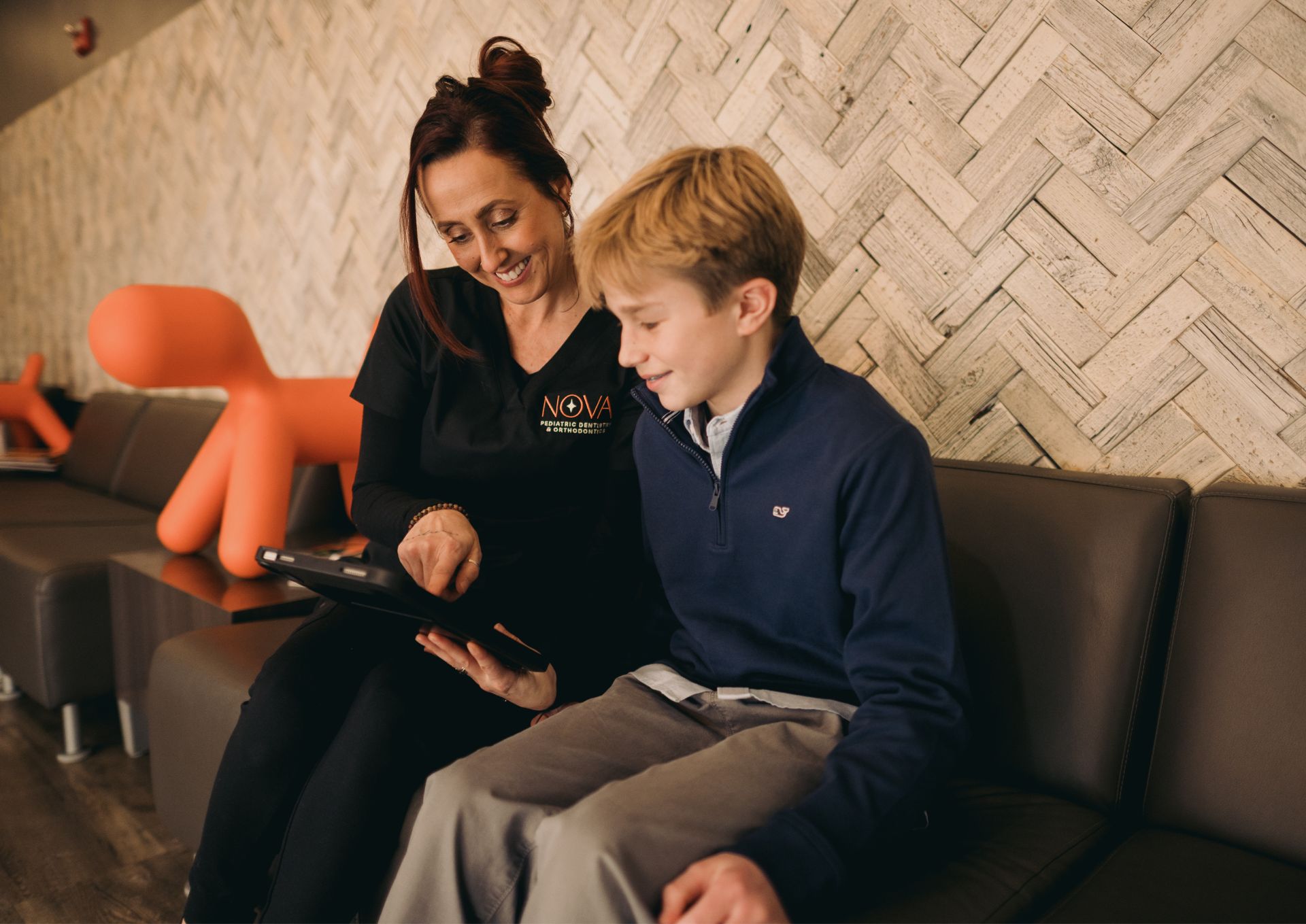 A woman in a Nova Pediatric Dentistry uniform showing a tablet to a smiling boy sitting on a bench in a dental office waiting area.