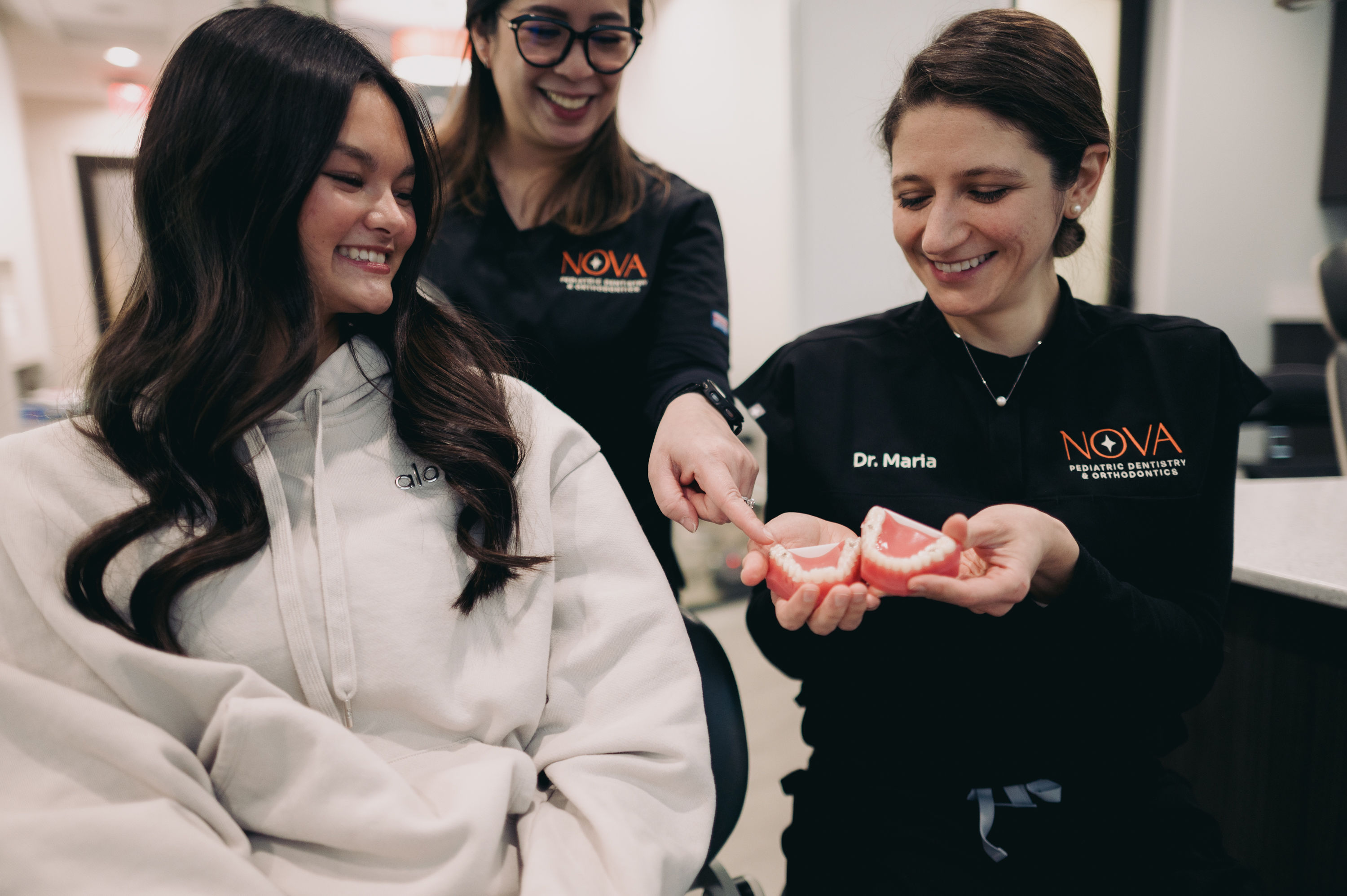 Dentist Dr. Maria smiling and holding dental models while explaining to a smiling female patient in a clinic.
