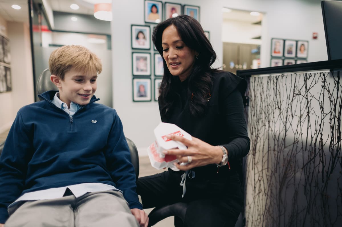 Young boy in dark jacket learning about orthodontics with dental professional showing aligner model