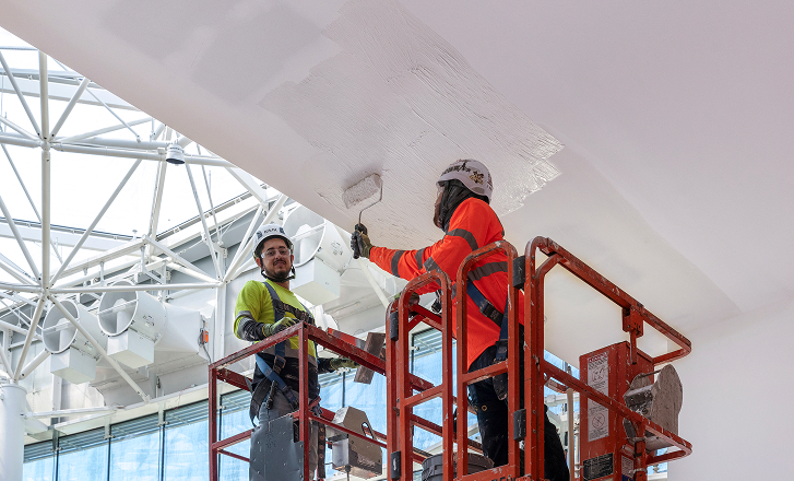 Two construction workers in safety gear paint a ceiling white while standing on a red scaffold inside a modern building with large windows and exposed beams.