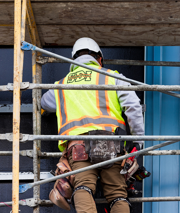Construction worker wearing a white hard hat and neon yellow safety vest working on scaffolding against a building wall.
