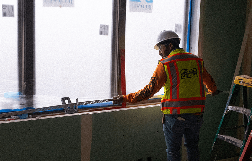 Construction worker in a hard hat and safety vest installing window framing inside a building under construction.