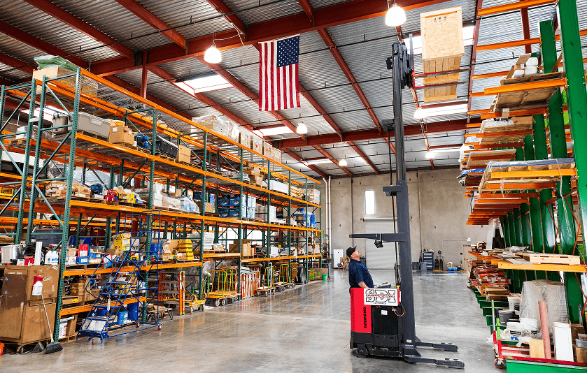 Worker operating a forklift in a large warehouse with shelves stocked with various materials and an American flag hanging from the ceiling.