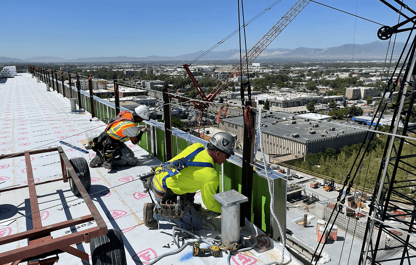 Two construction workers in safety gear working on the edge of a high building under a clear blue sky, with a cityscape and mountains in the background.