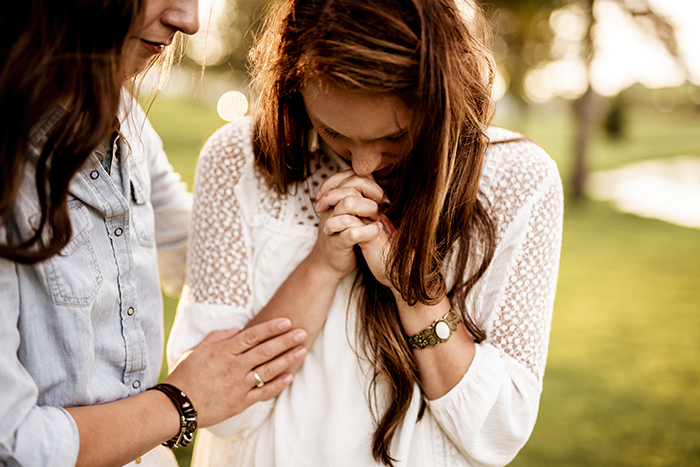Woman comforting another woman who is clasping her hands and looking down outdoors.