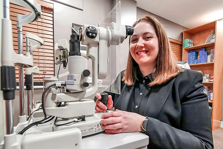 Smiling woman sitting beside ophthalmic equipment in an eye care clinic room.