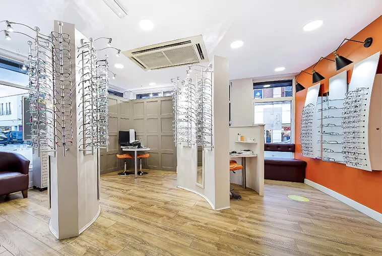 Interior of an eyewear store featuring multiple displays of eyeglasses on walls and freestanding racks with wooden flooring and seating areas.