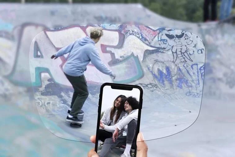 Person skateboarding in a graffiti-covered skate park viewed through a large eyeglass lens held in front, with a phone showing a selfie of two people sitting together.