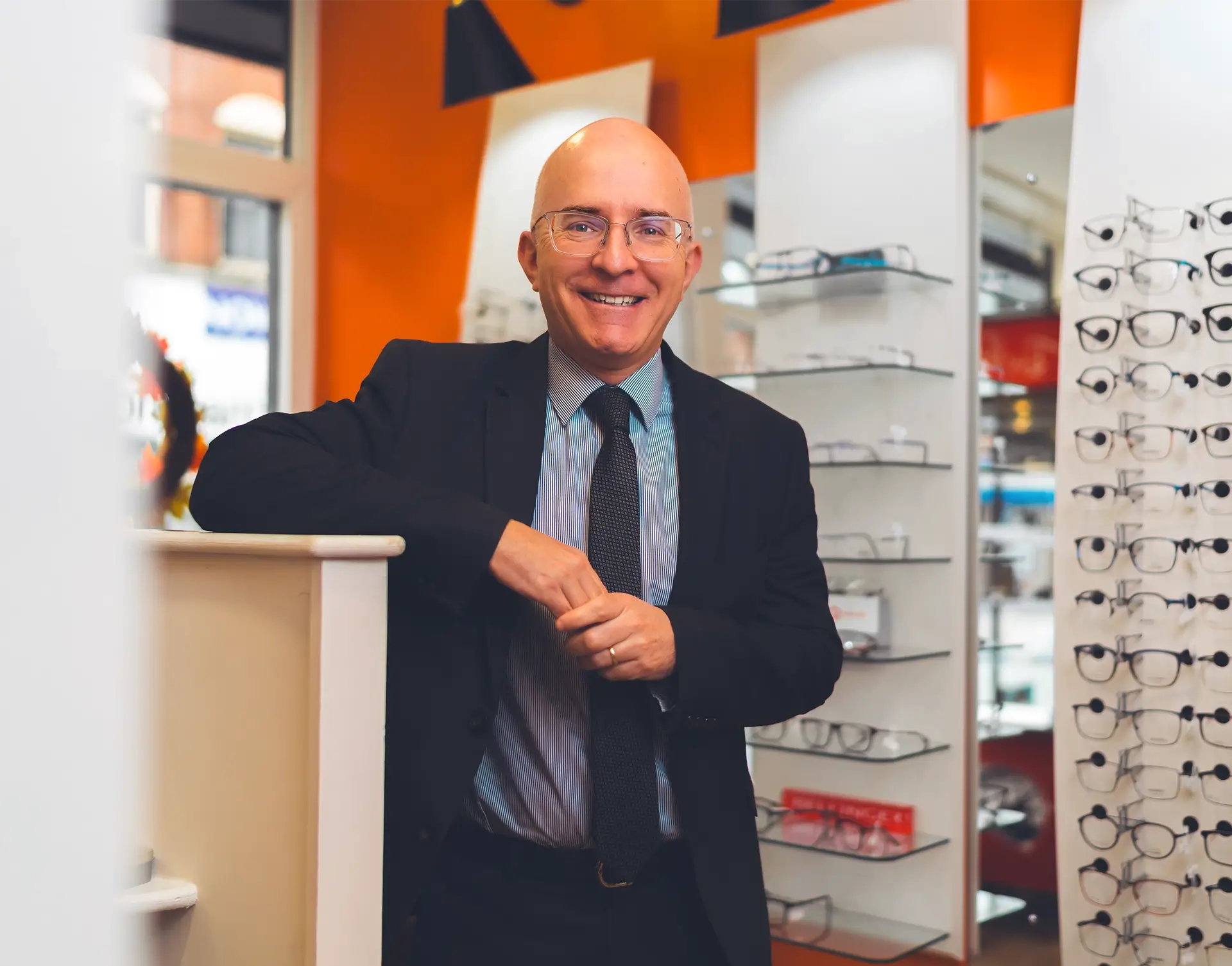 Smiling bald man wearing glasses and a suit standing in an eyewear store with glasses displayed on shelves behind him.