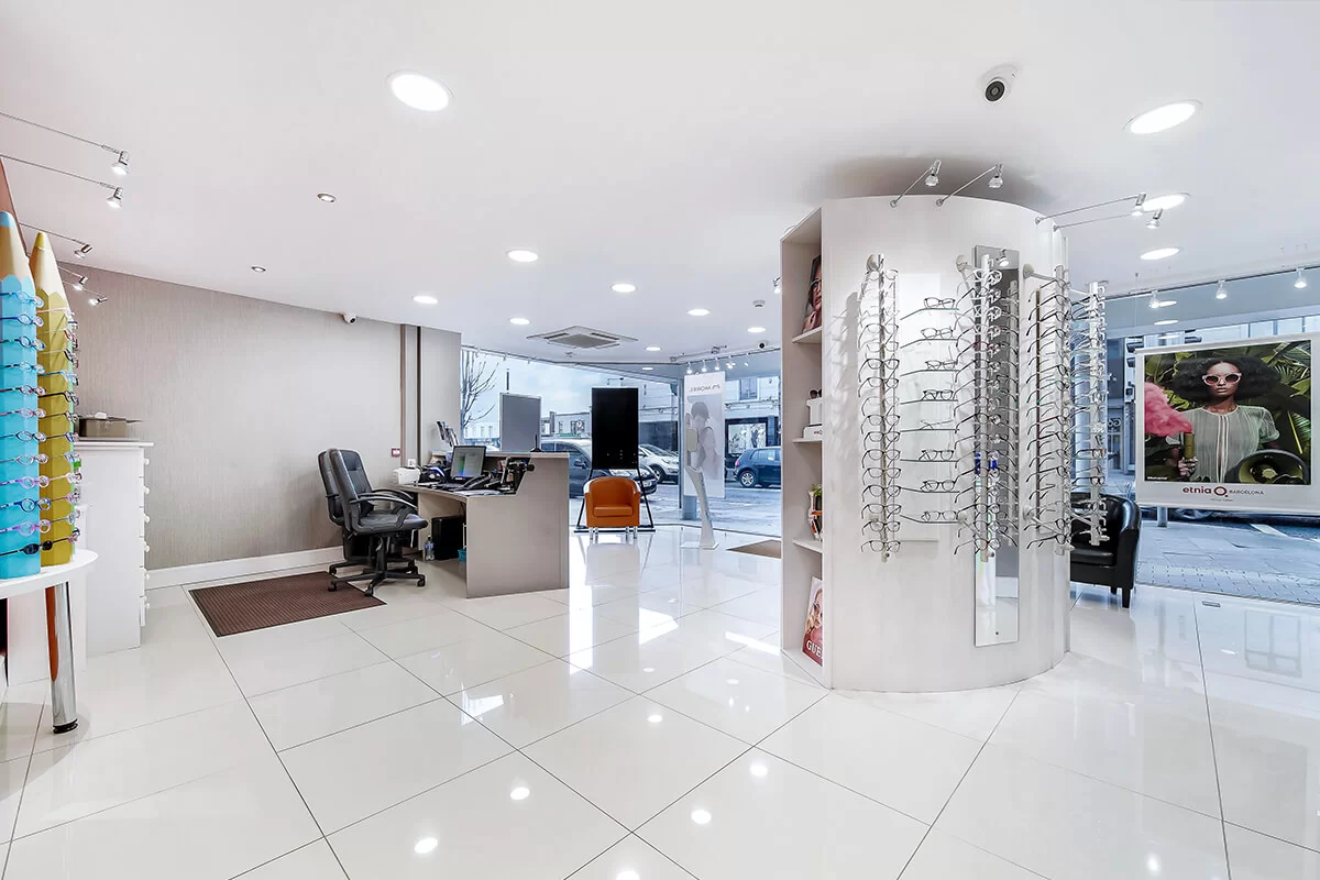 Bright, modern optician store interior with white tiled floor, eyeglass displays, office desks, and large windows showing a street view.