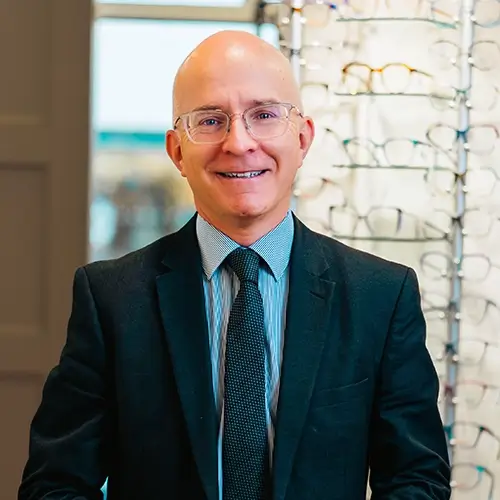 Smiling Mark Gormley wearing glasses, a dark suit, striped shirt, and tie in an indoor setting with eyeglasses displayed in the background.