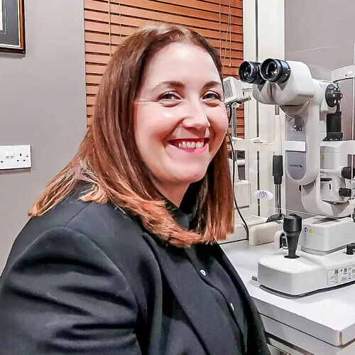 Smiling Roisin with brown hair sitting next to an ophthalmic examination device in an eye clinic.