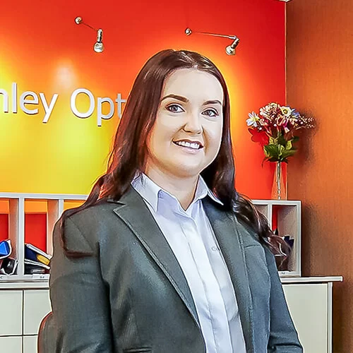 Gemma with long brown hair wearing a white shirt and gray blazer sitting and smiling in front of an orange-yellow wall with partial text and a vase of flowers on a cabinet.