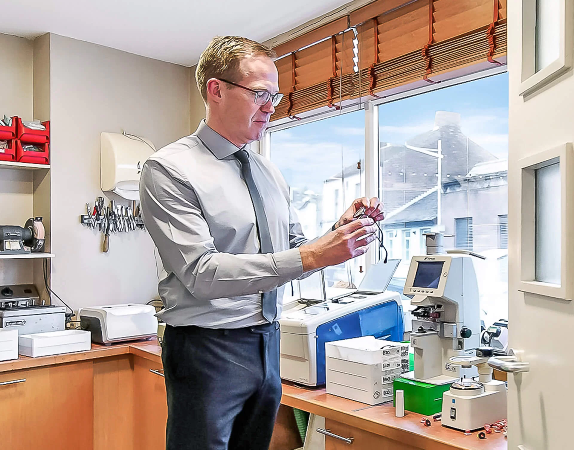 Man in a dress shirt and tie examining a pair of eyeglasses in an optical lab with various equipment and tools on the counters.