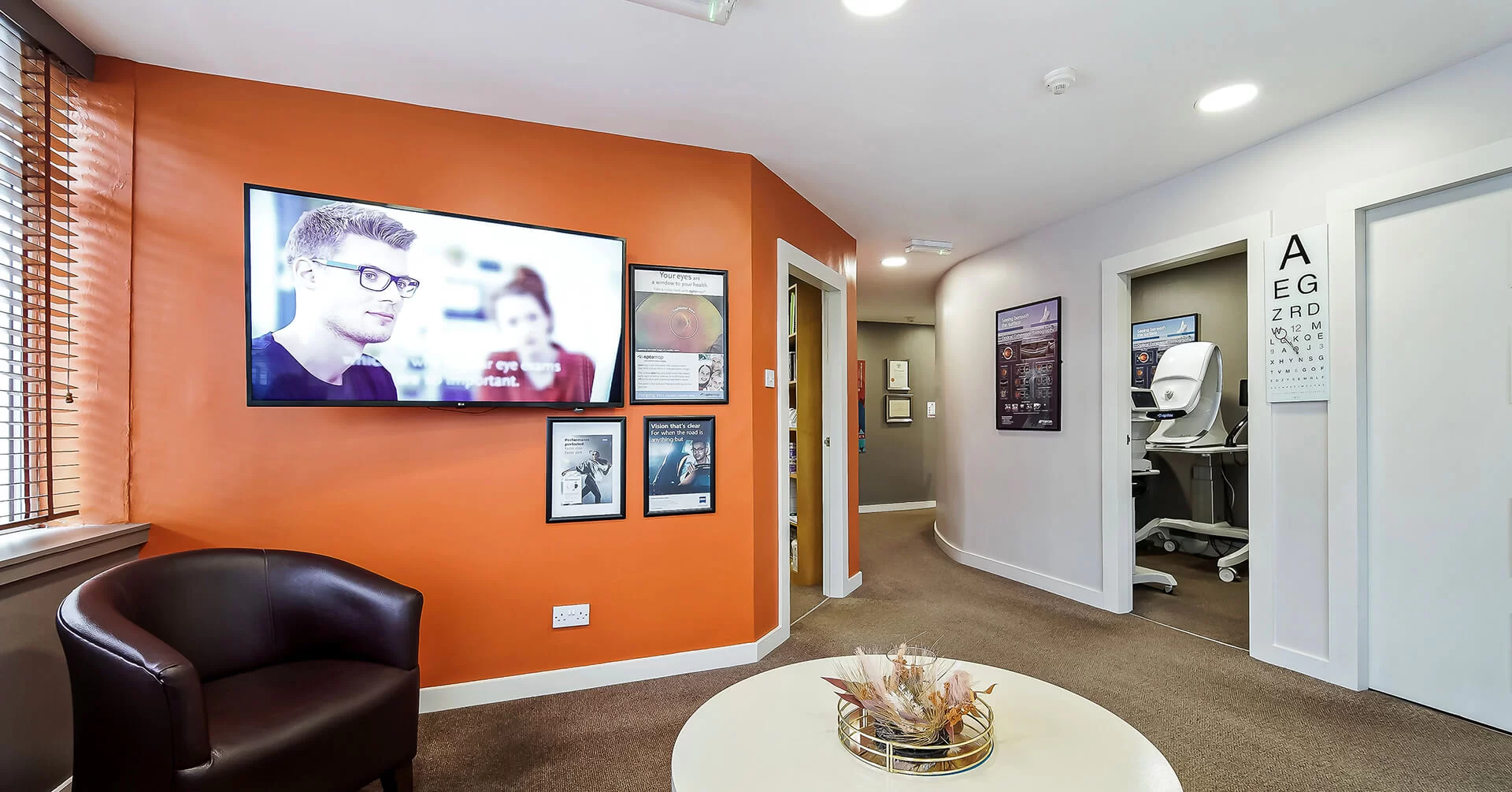 Modern optometry clinic waiting area with orange accent wall, mounted TV, eye charts, and diagnostic equipment visible through an open door.