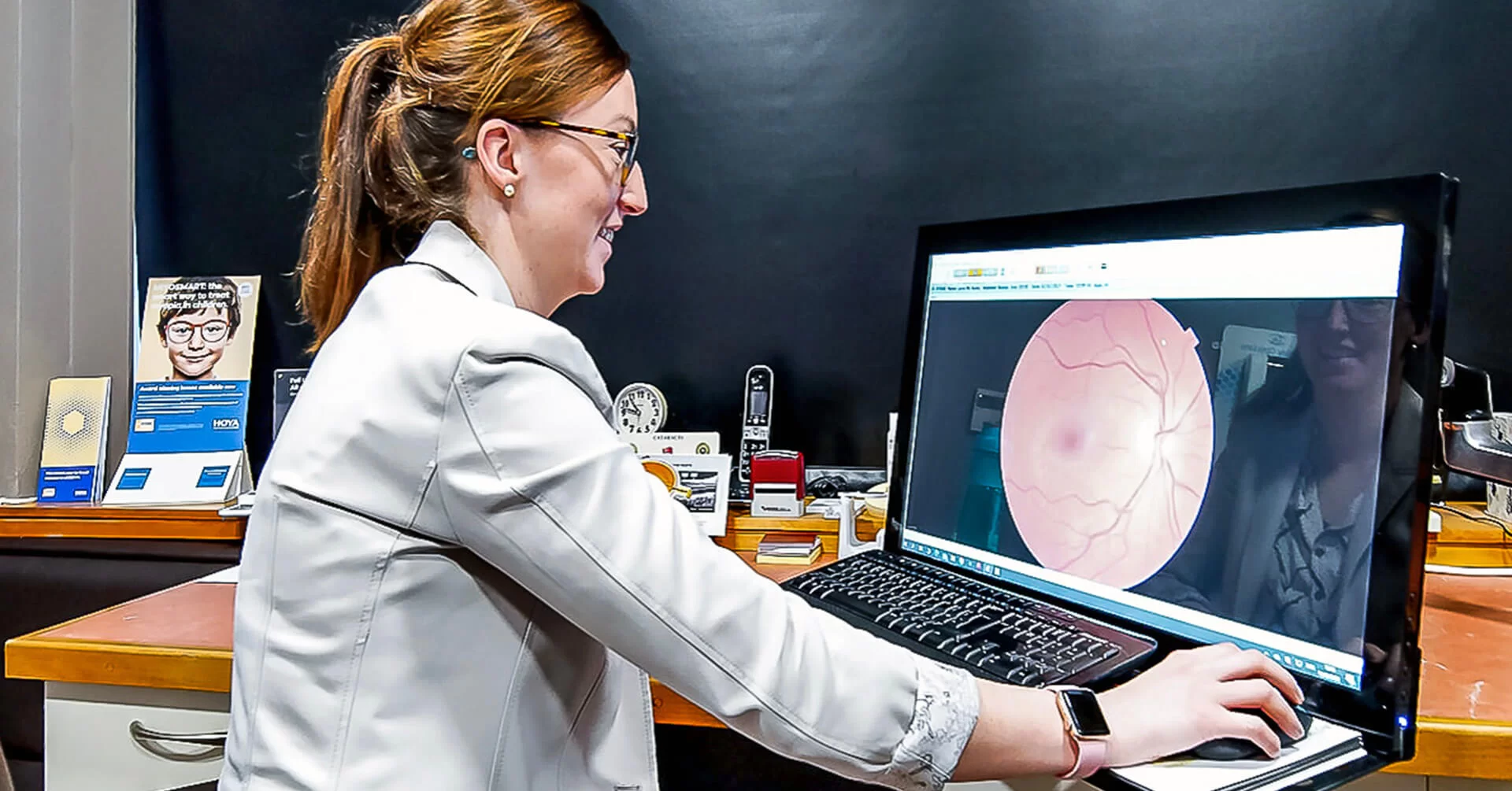 Optician with glasses reviewing a detailed eye retina scan on a computer screen in an office setting.