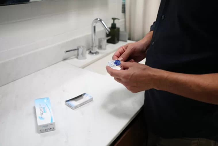 Person holding a contact lens case near a bathroom sink with contact lens boxes on the countertop.