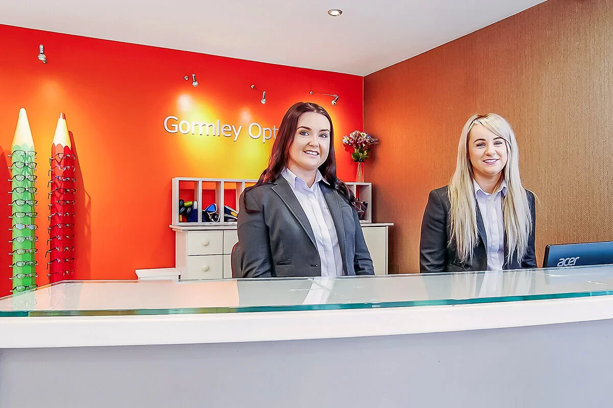Two women in business attire smiling behind a glass counter in an optician office with a bright orange wall and eyeglass displays.