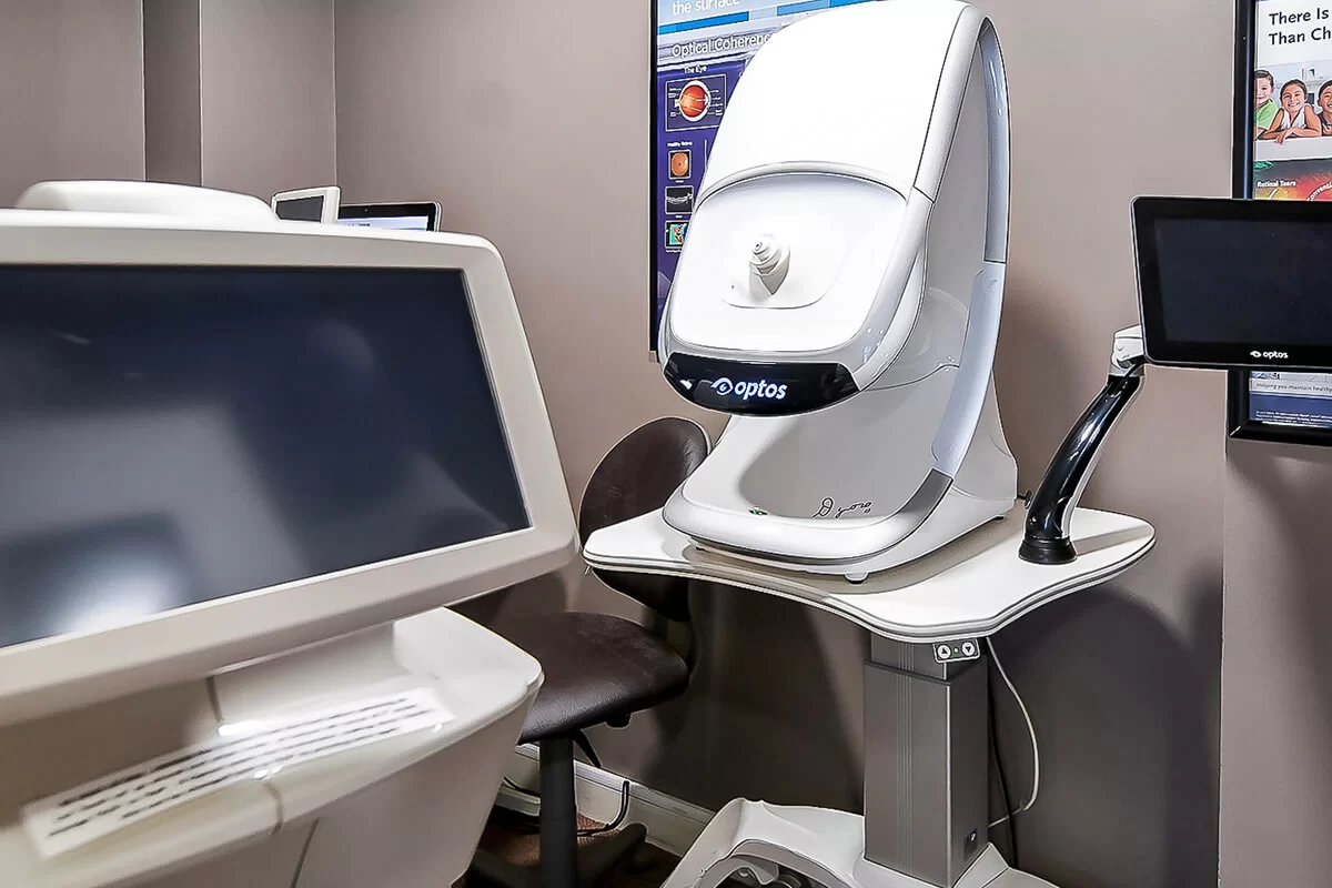 Optos retinal imaging machine in an eye exam room with two black monitors and a chair.