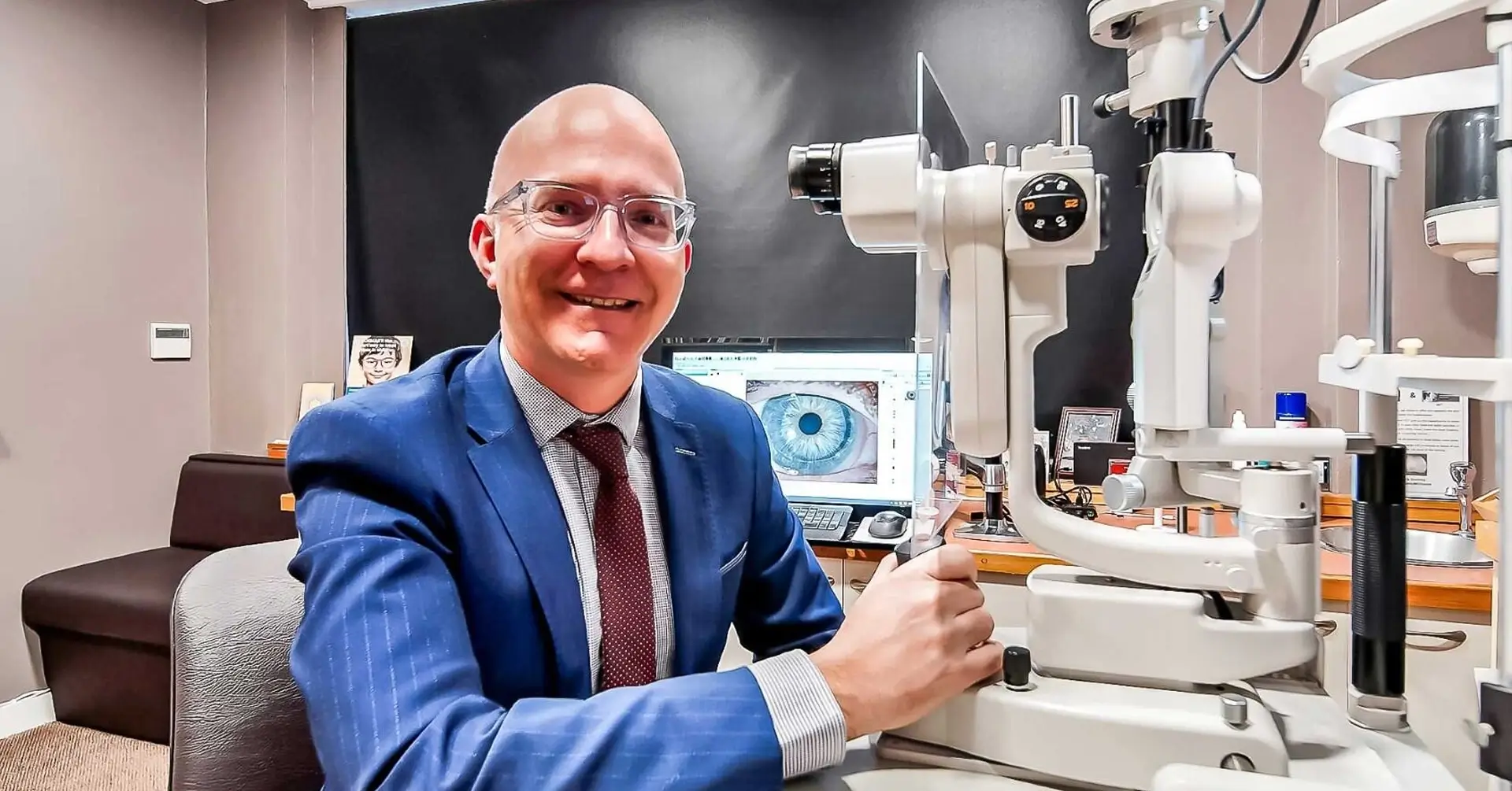 Smiling man in a blue suit sitting at an eye examination machine in an optometrist office.
