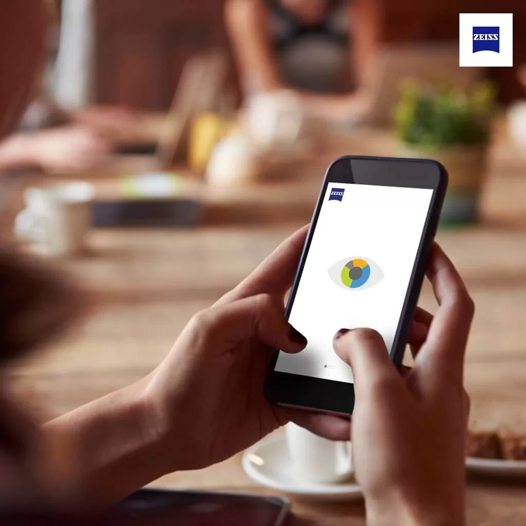 Person holding a smartphone displaying a colorful eye icon and Zeiss logo on the screen in a coffee shop setting.