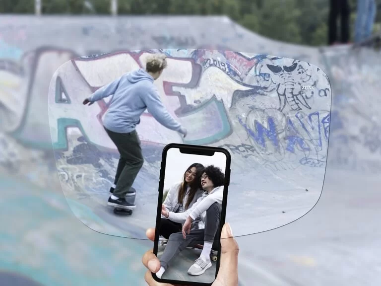 A hand holding a smartphone displays a photo of two smiling people sitting, superimposed over a skateboarder in a graffiti-covered skatepark.