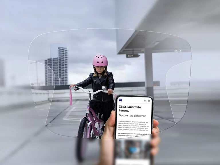View through ZEISS SmartLife lenses showing a child wearing a pink helmet riding a bike, with a hand holding a phone displaying information about ZEISS SmartLife lenses.