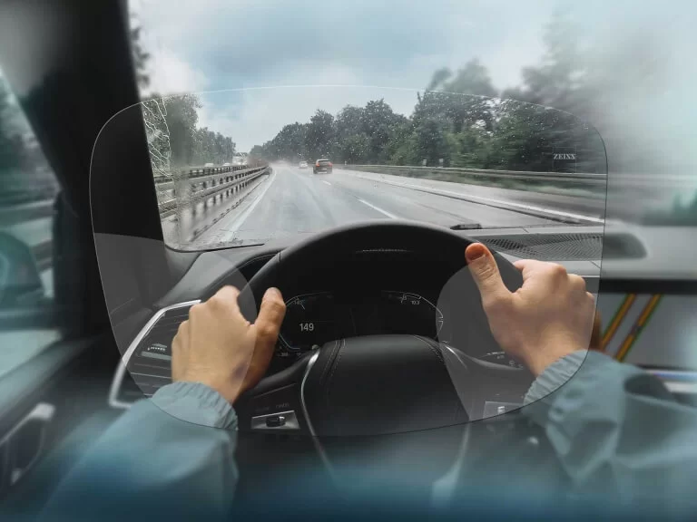 View from inside a car showing hands on the steering wheel and a clear windshield with raindrops while driving on a wet road.