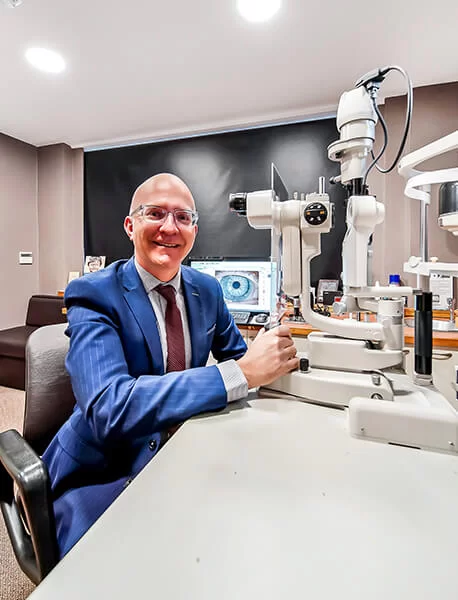 Smiling male optometrist in a blue suit sitting next to eye examination equipment in an eye care clinic.