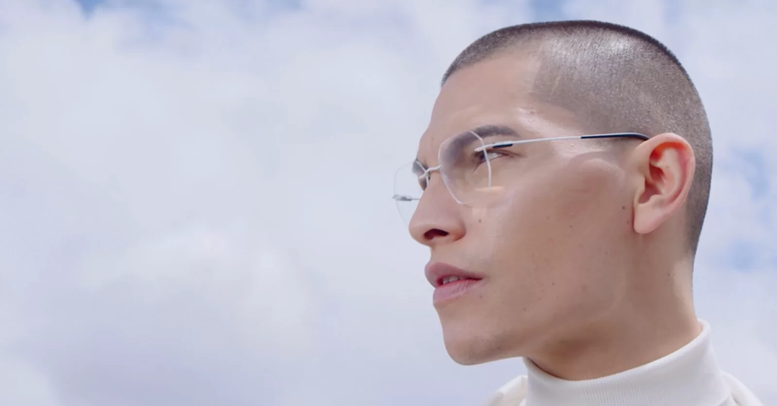 Profile of a man with short hair wearing thin-framed clear eyeglasses and a white turtleneck against a cloudy sky.