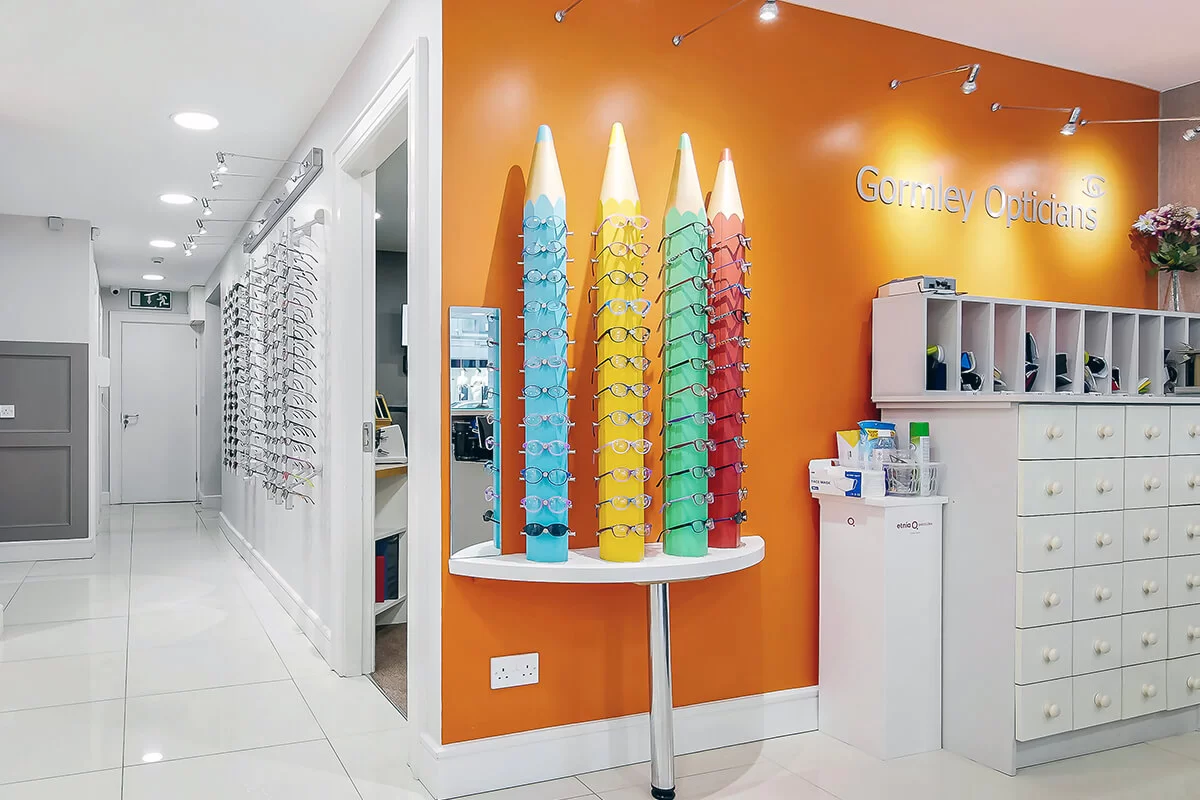 Interior of Gormley Opticians with colorful pencil-shaped eyeglass displays and white cabinets under bright lighting.