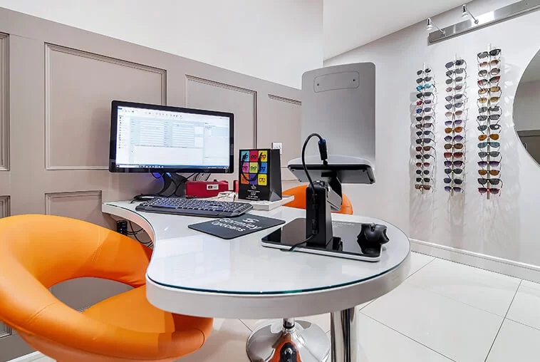 Modern eyewear store interior with a curved white desk, computer setup, orange chairs, and sunglasses displayed on the wall.
