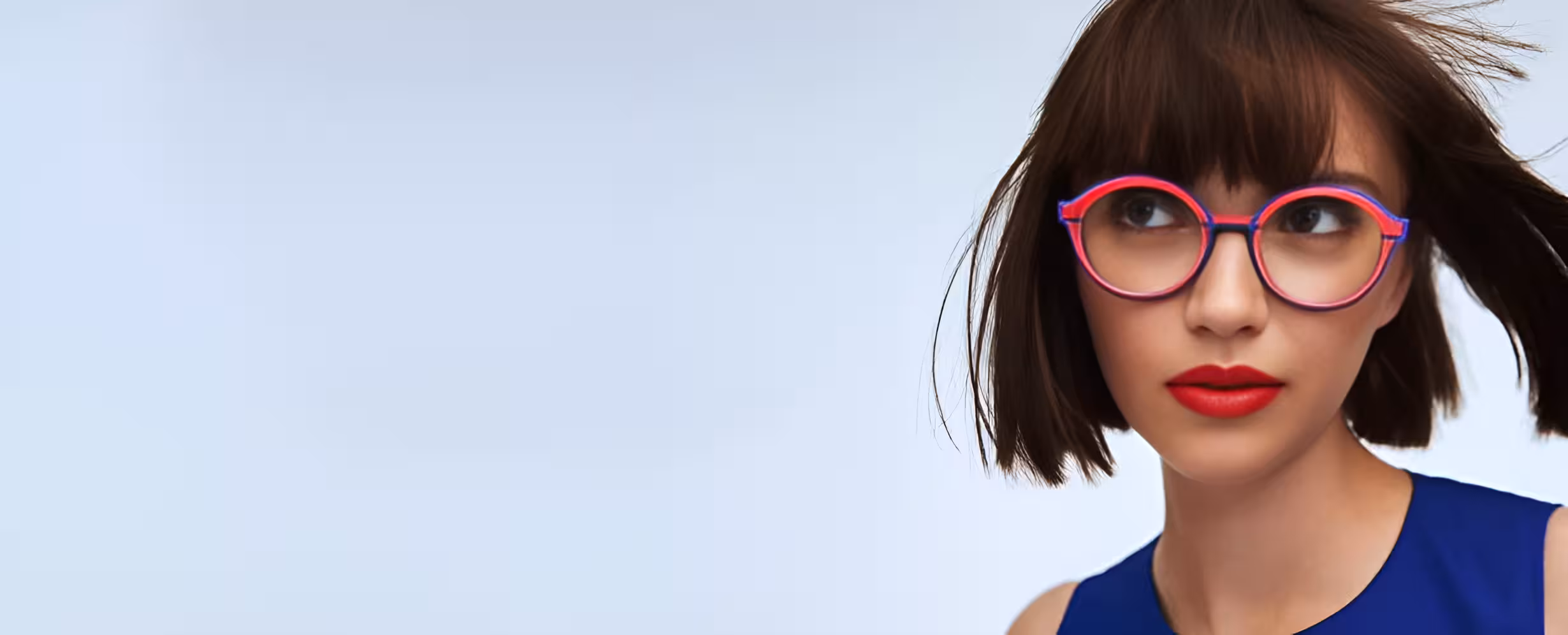 Young woman with short brown hair wearing red lipstick and round purple glasses looking to the side against a light blue background.