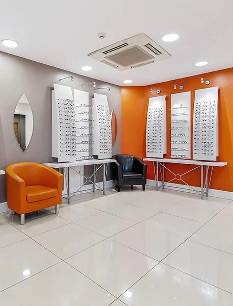 Modern eyeglass store interior with display panels holding multiple glasses, orange and black chairs, and bright lighting.